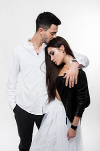 Beautiful brunette girl in a white tulle skirt in a black bra and black leather jacket with guy in trousers and white t-shirt are hugging on the white background in the studio picture